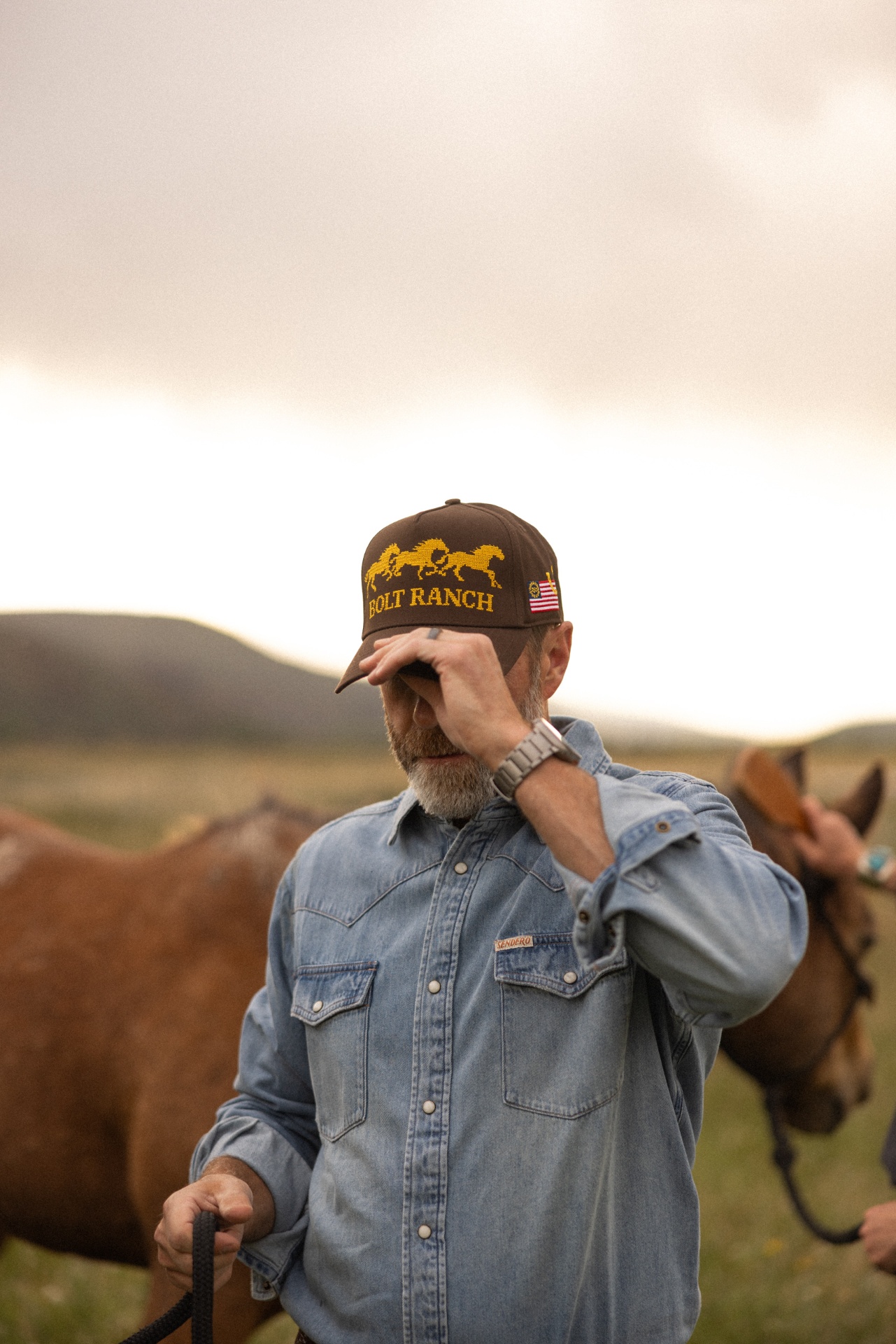 Cowboy in a Bolt Ranch cap adjusting the brim while standing beside horses